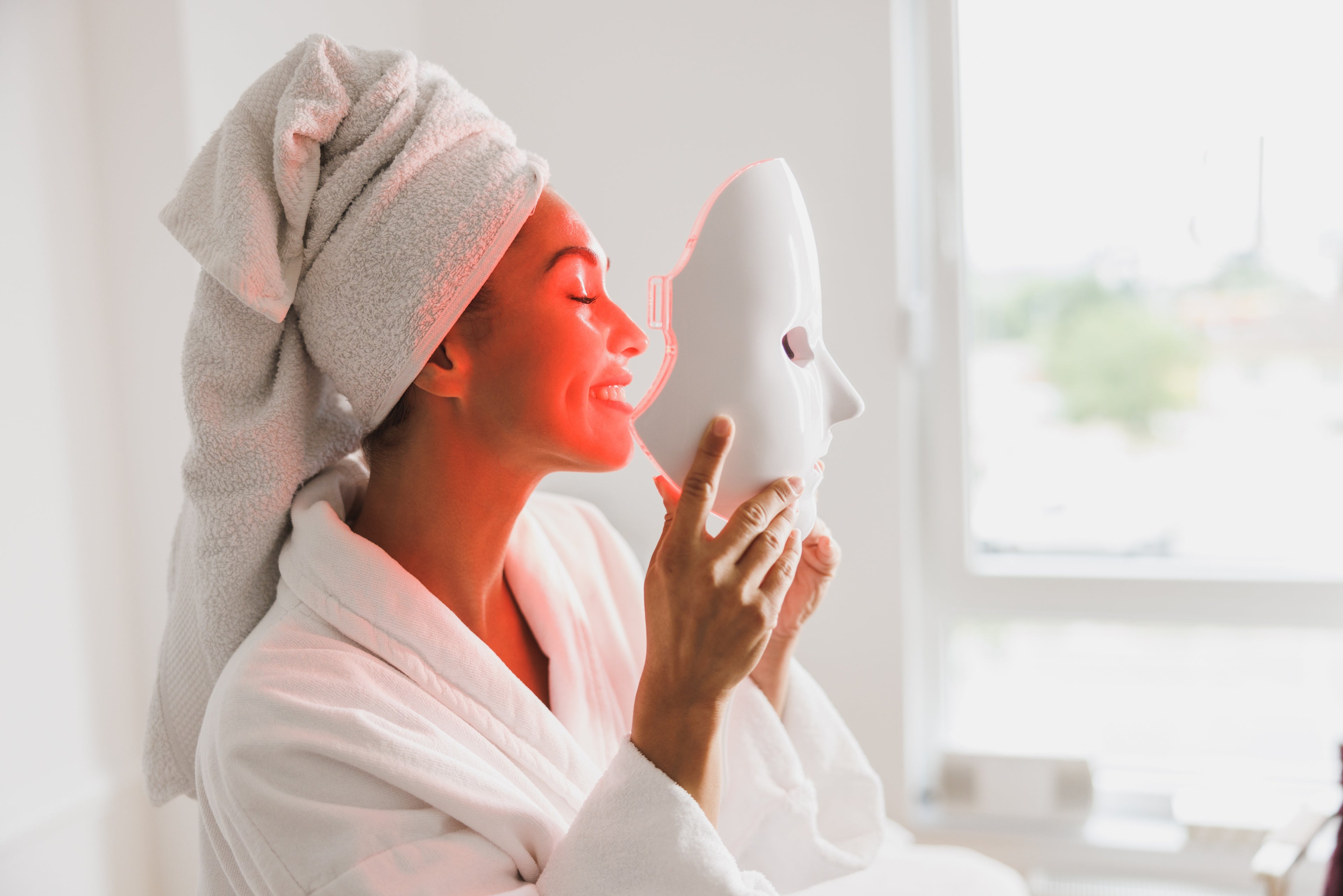 Woman in a bathrobe holding a white face mask with a towel on her head, sitting by a window.