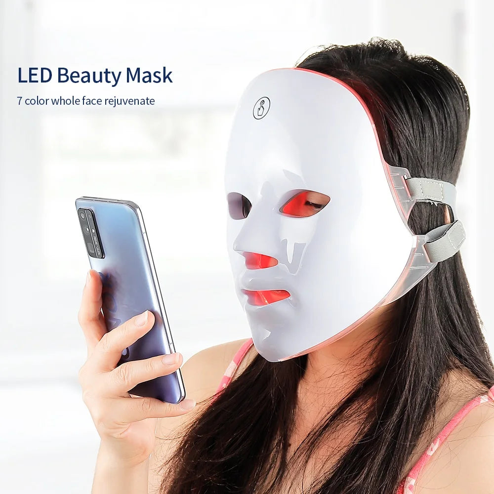 Woman wearing an LED beauty mask holding a phone with a white background